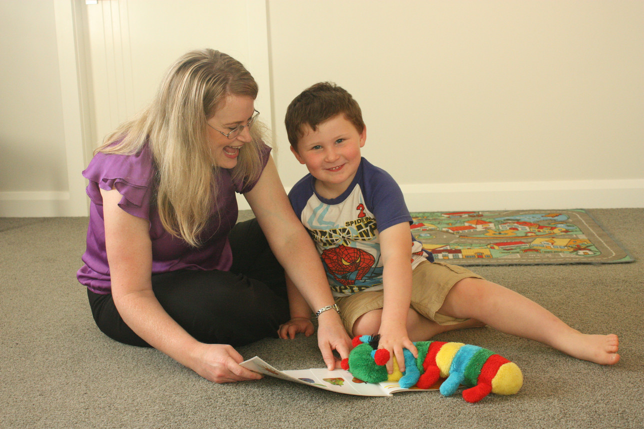 Sarah and a young boy sitting on the floor sharing a picture book, a caterpillar toy beside them